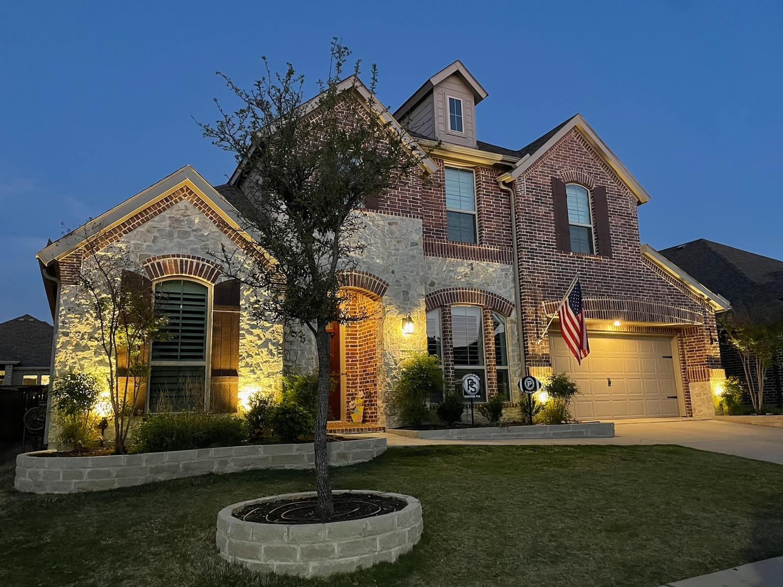 Two-story stone house at dusk with lit windows, a front yard tree, and an American flag by the garage.