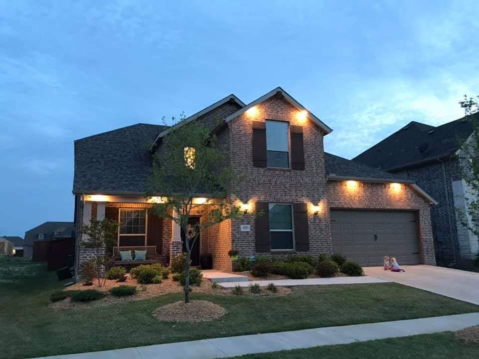 Two-story brick house with warm exterior lights at dusk in a suburban neighborhood