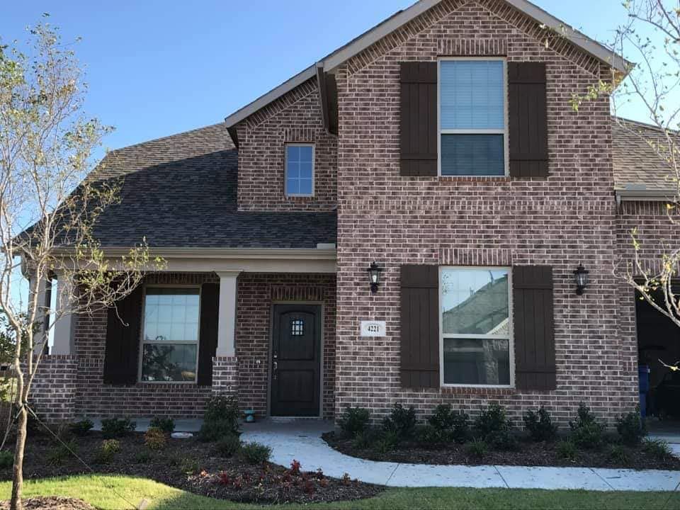 Front view of a two-story brick house with dark shutters, a front porch, and landscaped yard.