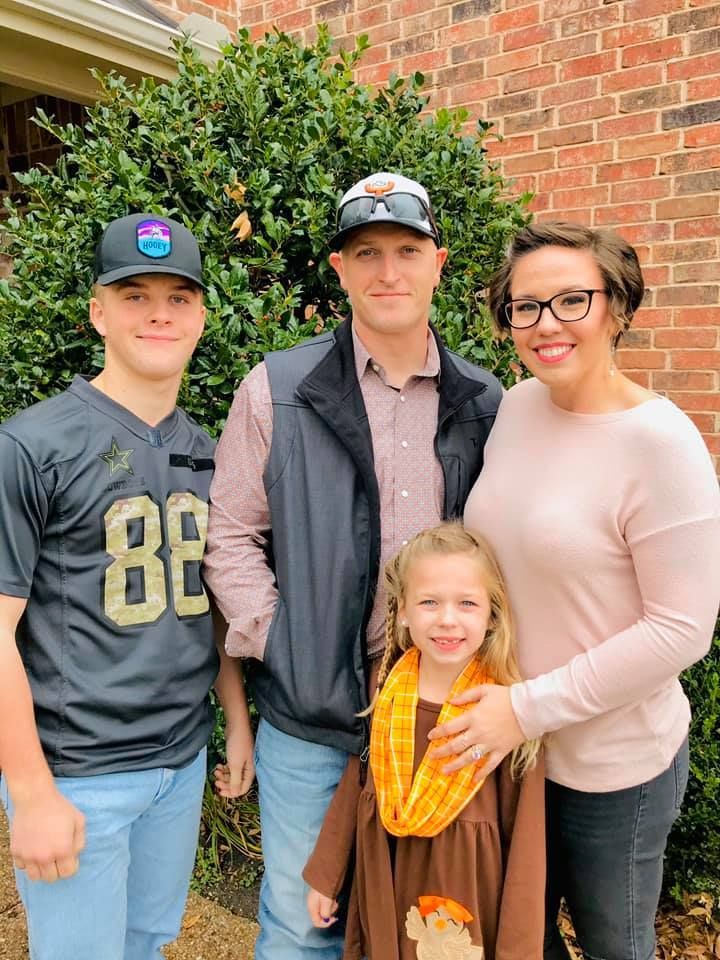 Four people standing outdoors in front of a brick wall, smiling for a family photo.