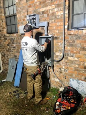 Electrician working on an outdoor wall-mounted electrical panel beside a brick building.