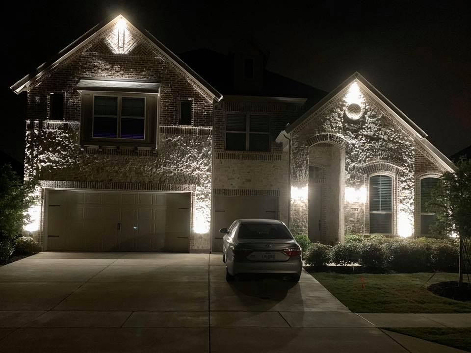 Nighttime view of a lit two-story brick house with a garage and a car parked in the driveway.