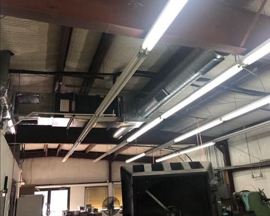 Industrial warehouse ceiling with exposed beams, ducts, and bright fluorescent lights above open floor equipment.
