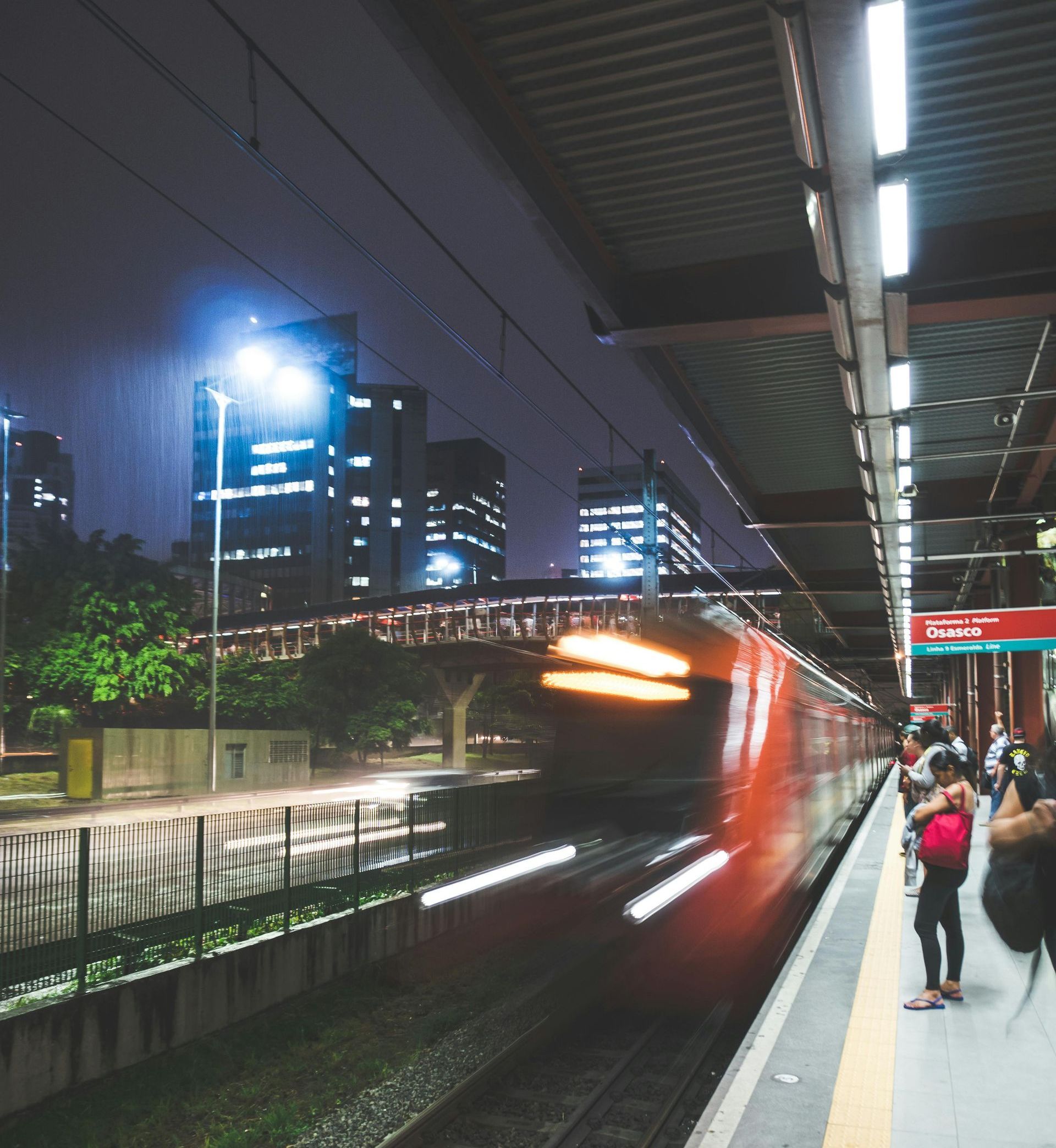 Tren rojo borroso que pasa a toda velocidad por una estación iluminada de noche con pasajeros en el andén.