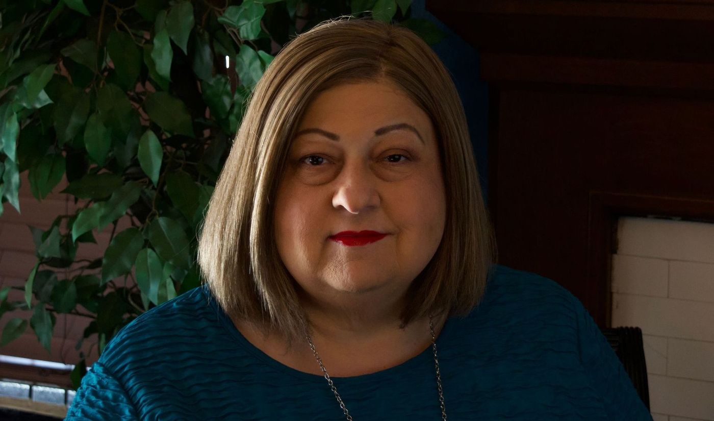 A woman in a blue sweater and red lipstick is standing in front of a fireplace.