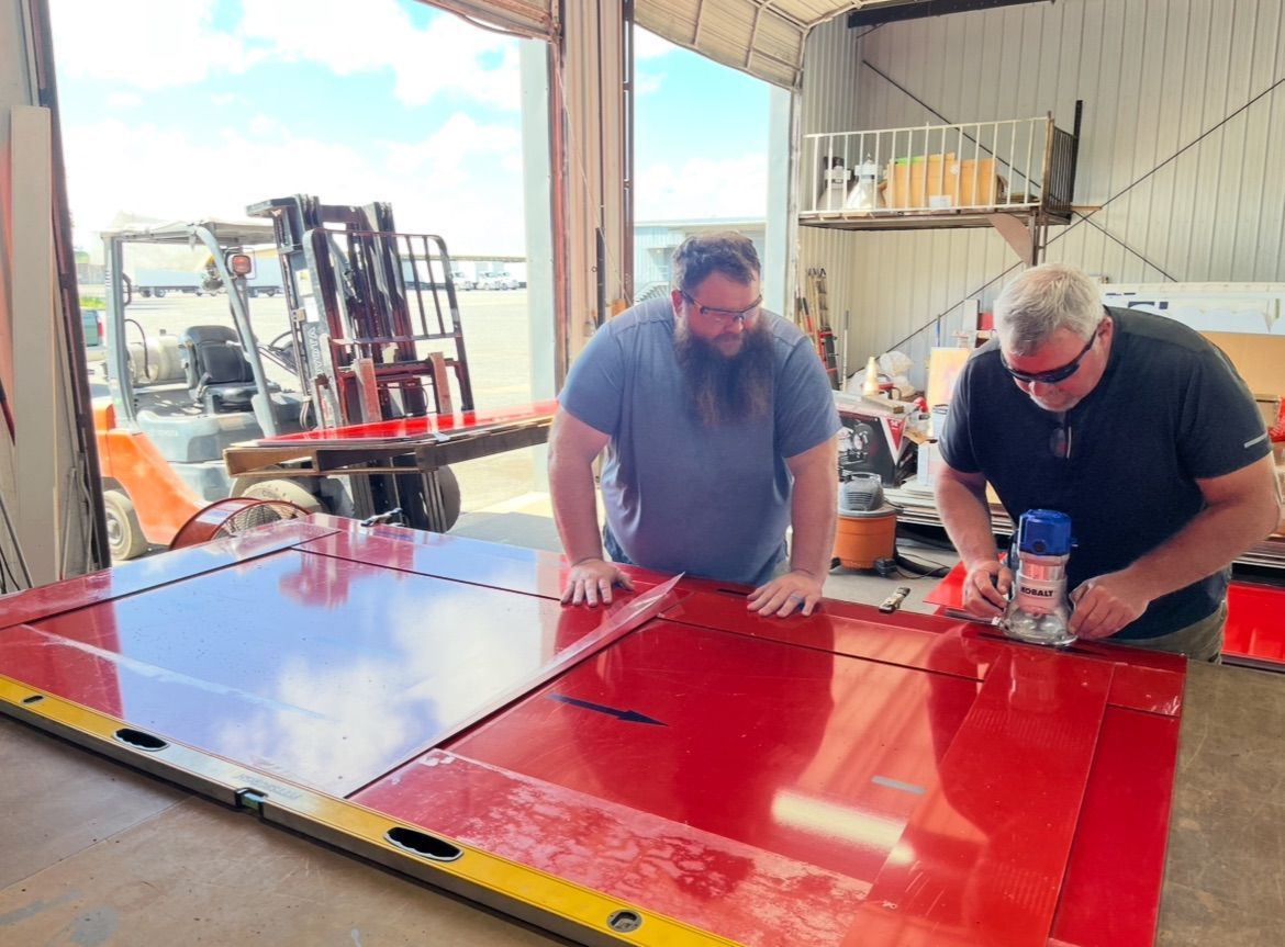Two men work on a red metal door in a workshop. One uses a router; the other watches. A forklift is in the background.