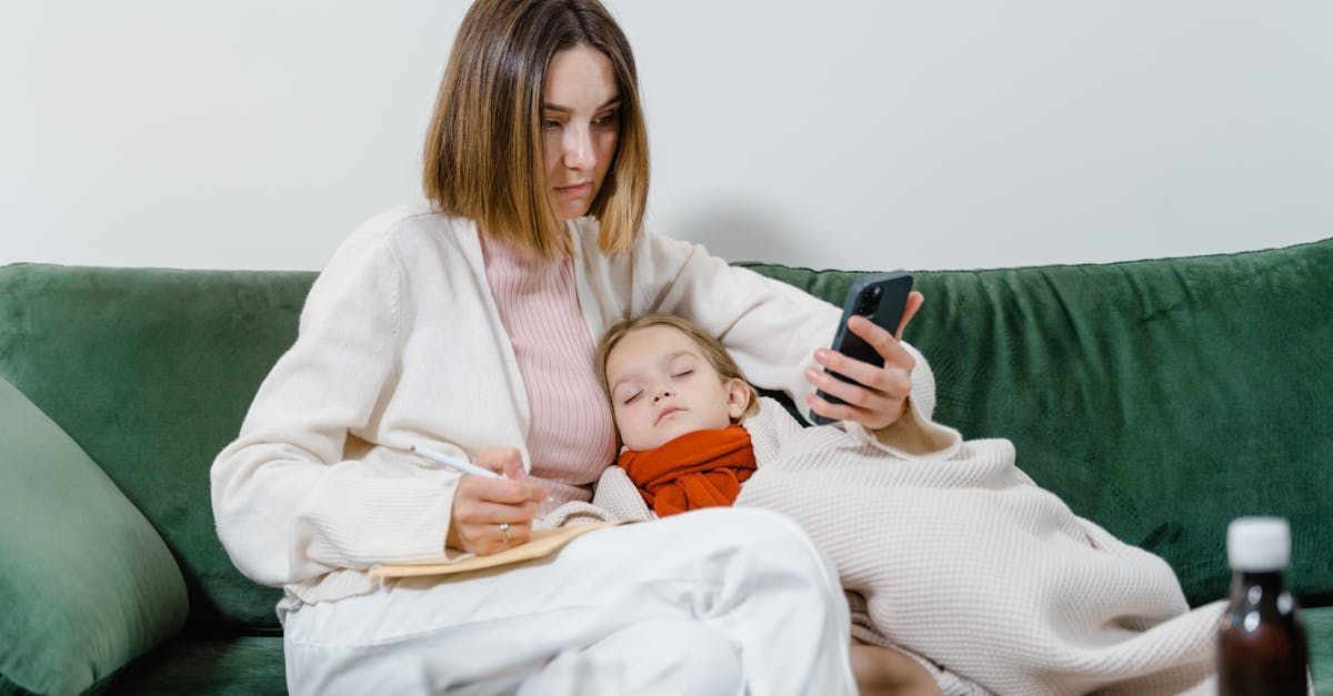 A woman is sitting on a couch with a child wrapped in a blanket and using a cell phone.