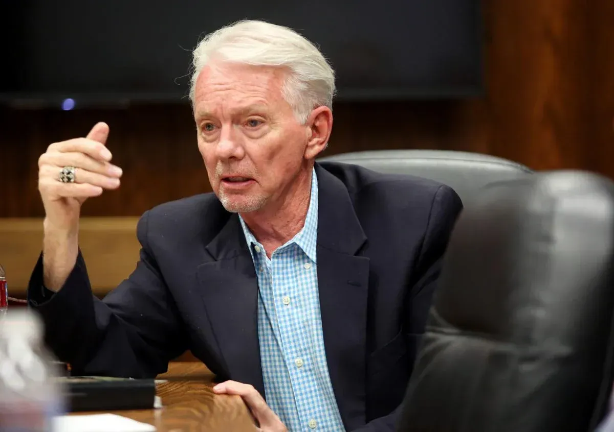 A person in a blue blazer and light blue patterned shirt sits at a table, gesturing while speaking during a meeting.