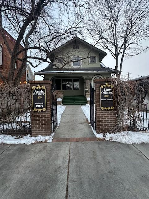 A path leads to an old house with a green porch between two brick pillars holding signs for 