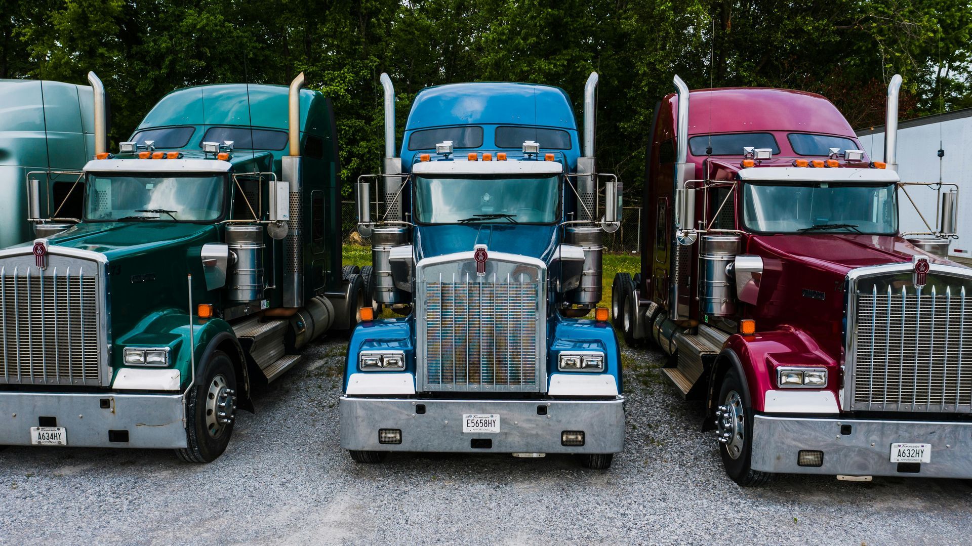 Three Kenworth semi-trucks in green, blue, and maroon parked side-by-side on a gravel lot.