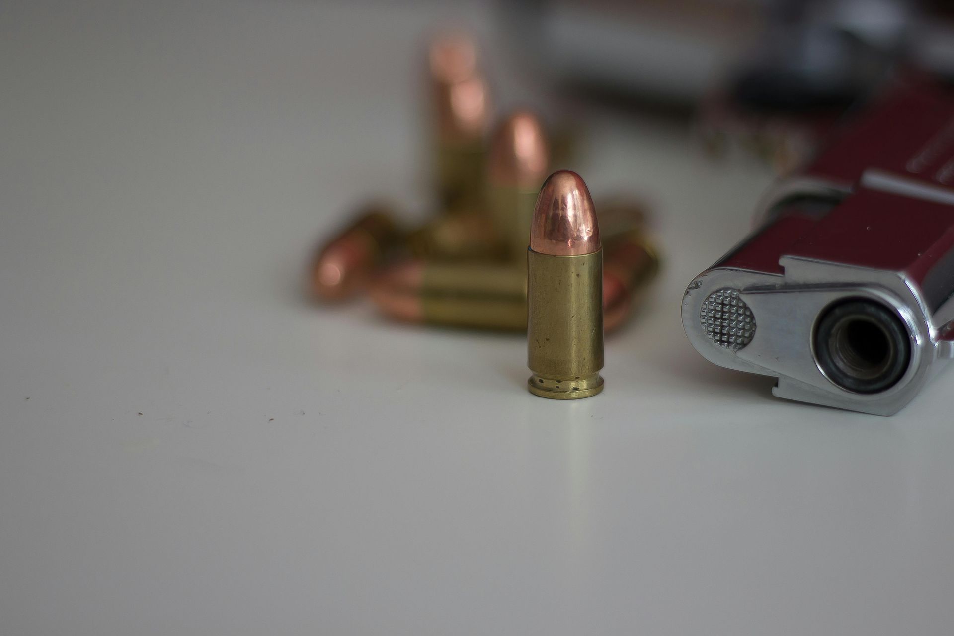 A single brass bullet stands upright on a white surface next to a firearm, with several blurred bullets in the background.