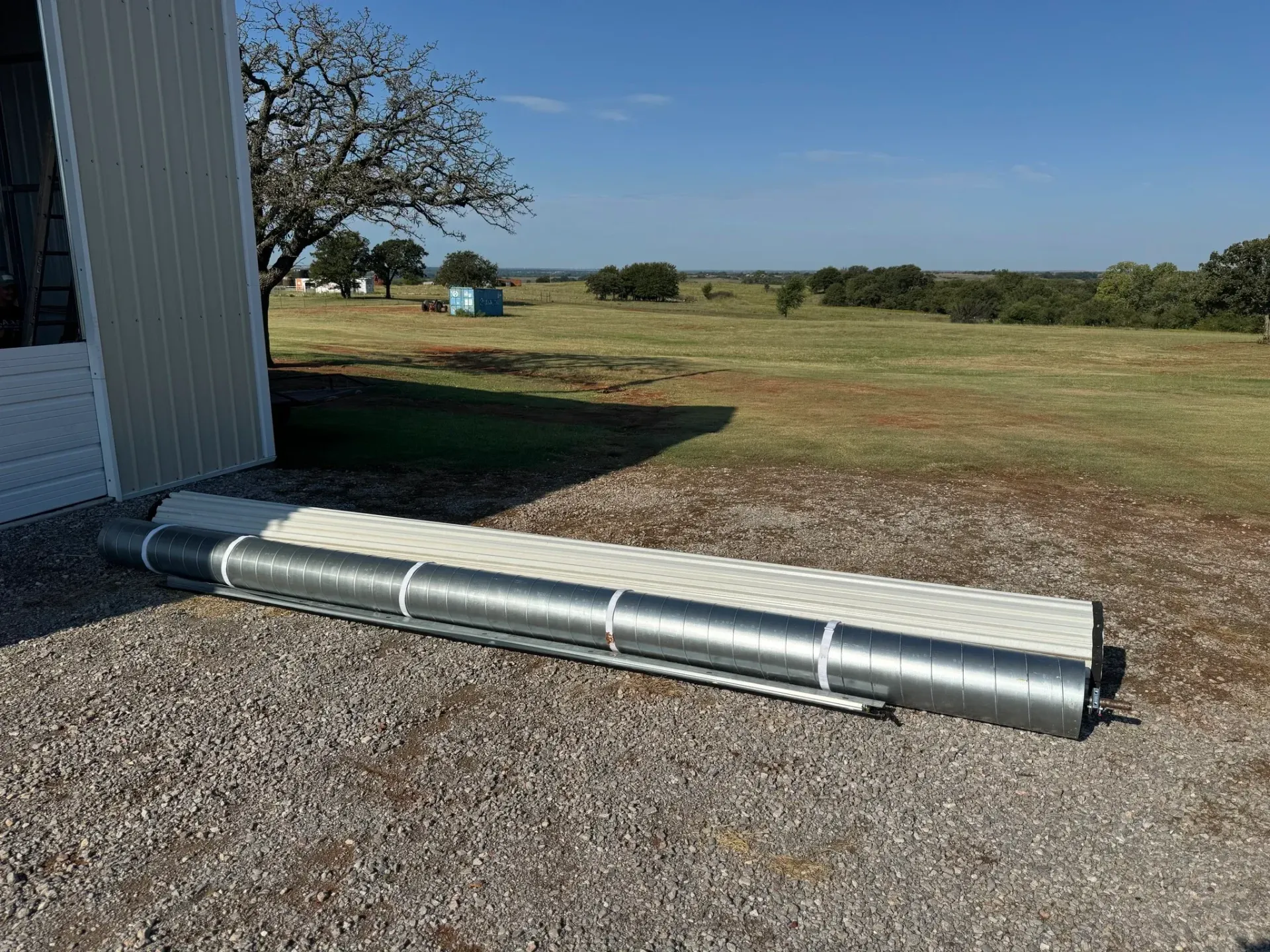 A rolled-up metal and white structure on gravel near a white building with a rural landscape in the background.