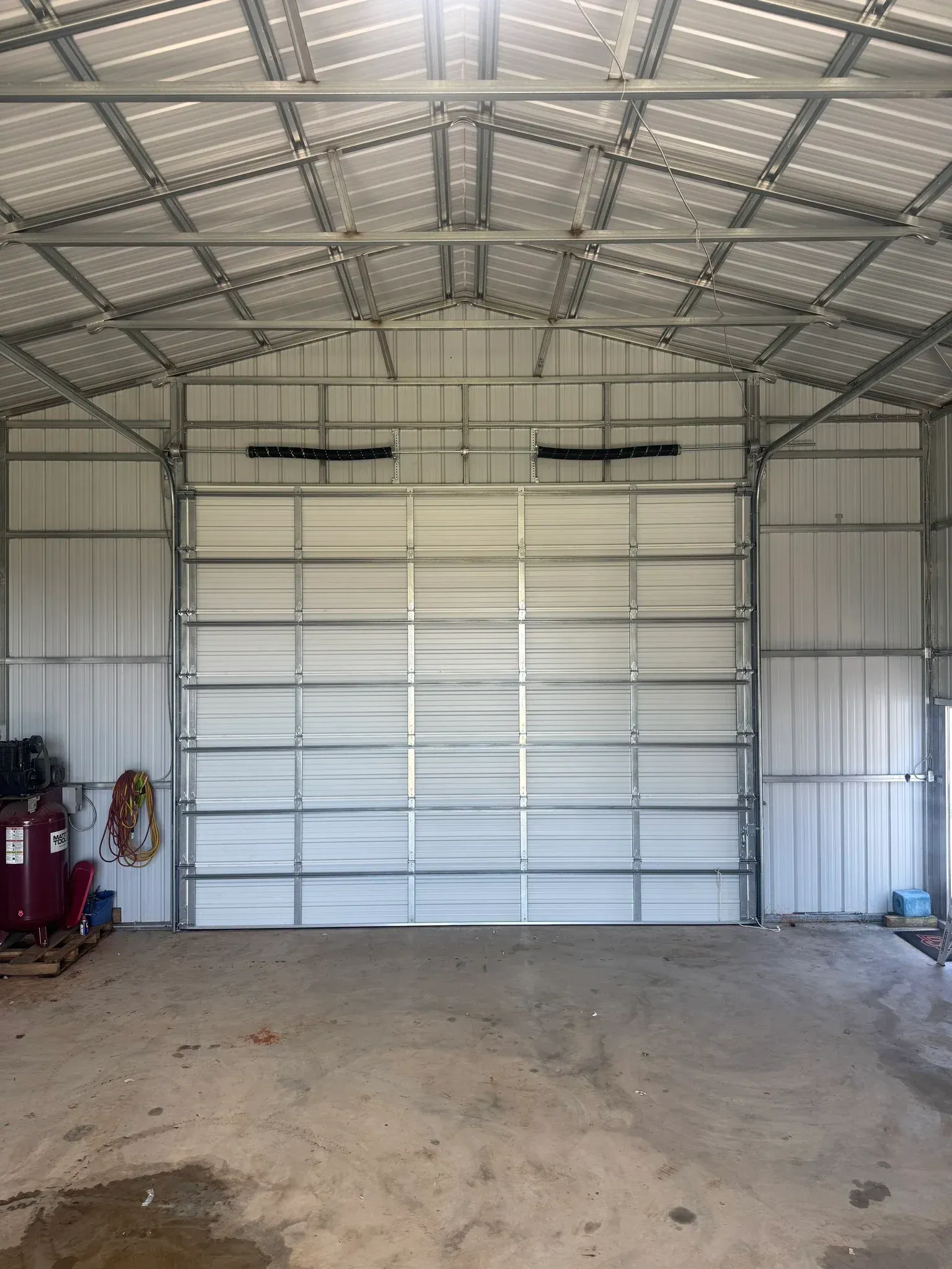 Inside of a metal-framed workshop, a closed garage door dominates the view; with exposed metal structure throughout the interior.