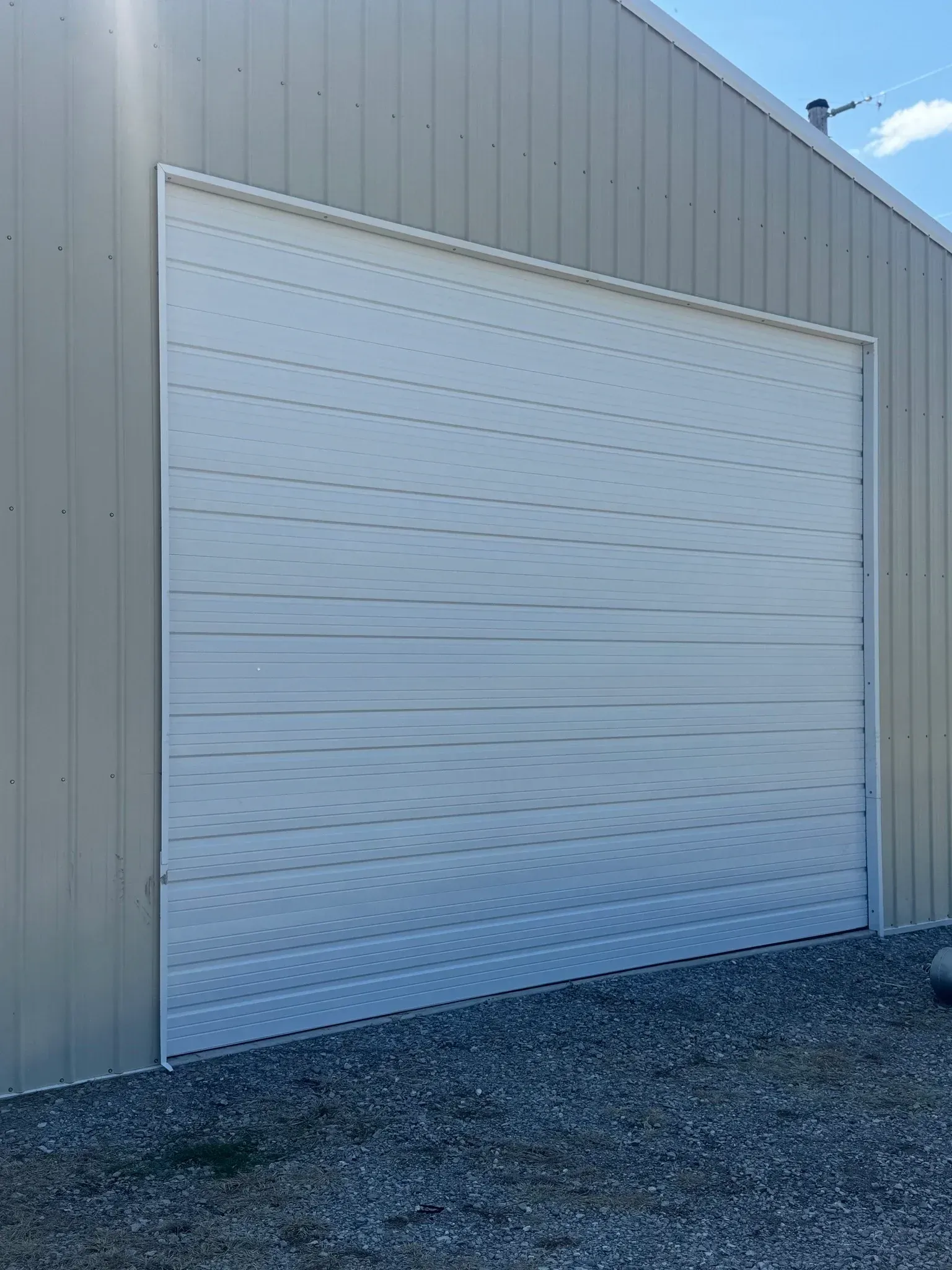 White overhead garage door on a tan metal building with a gravel base, under a blue sky.