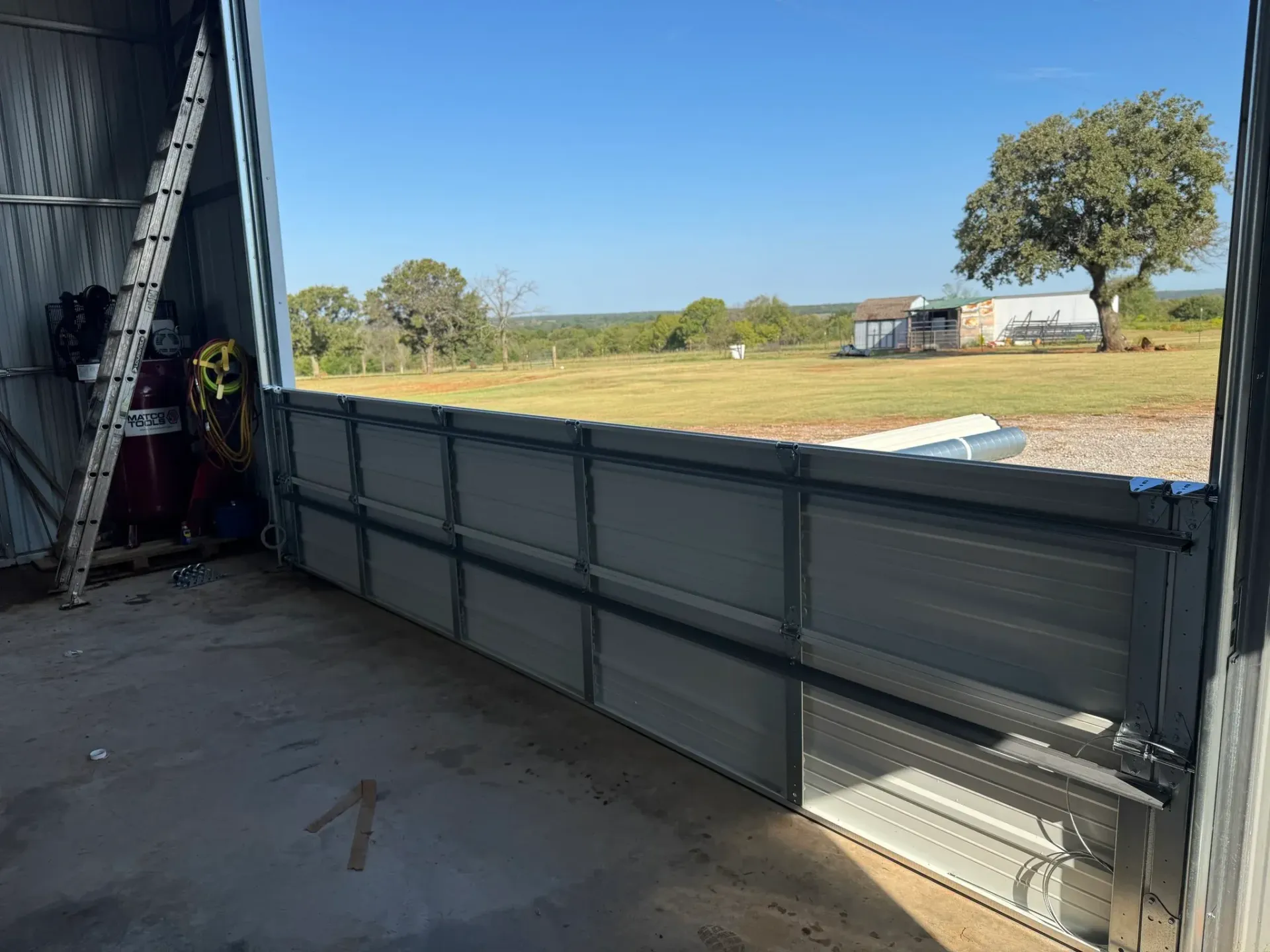 View from inside a metal structure, looking out at a field, house, and tree under a blue sky.
