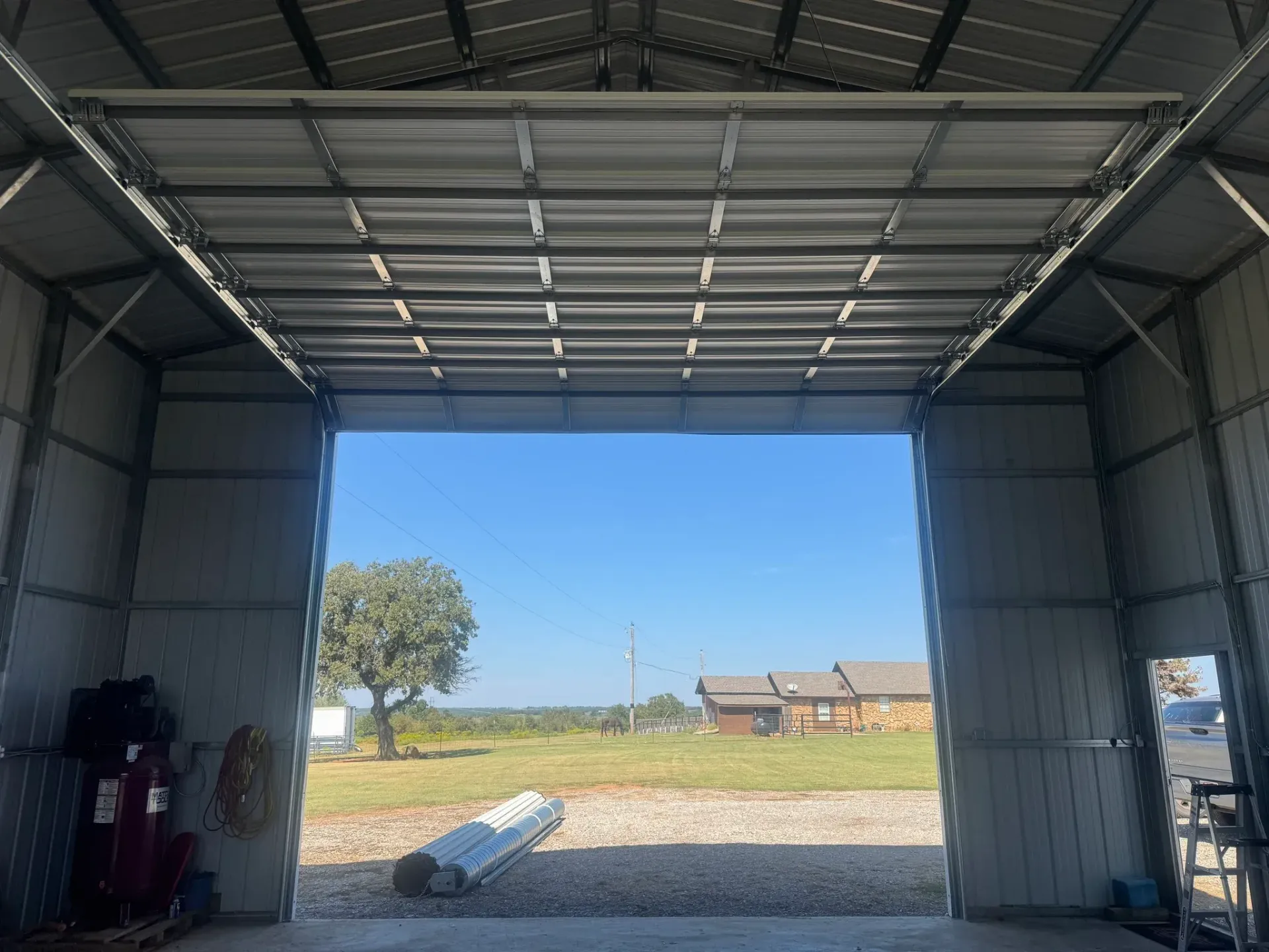Inside of a metal building, view through open door of a field with trees and blue sky.