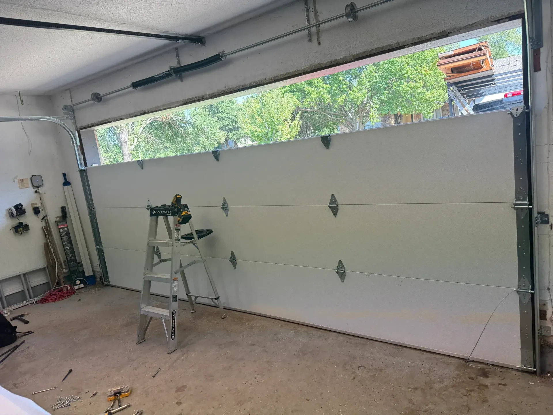 Garage door partially open; ladder inside. White door panels, visible outside greenery.