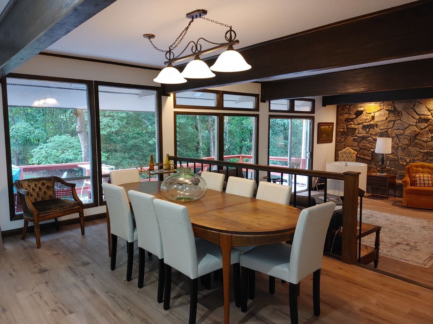 Dining room with wooden table, white chairs, hanging lights, and large windows overlooking greenery