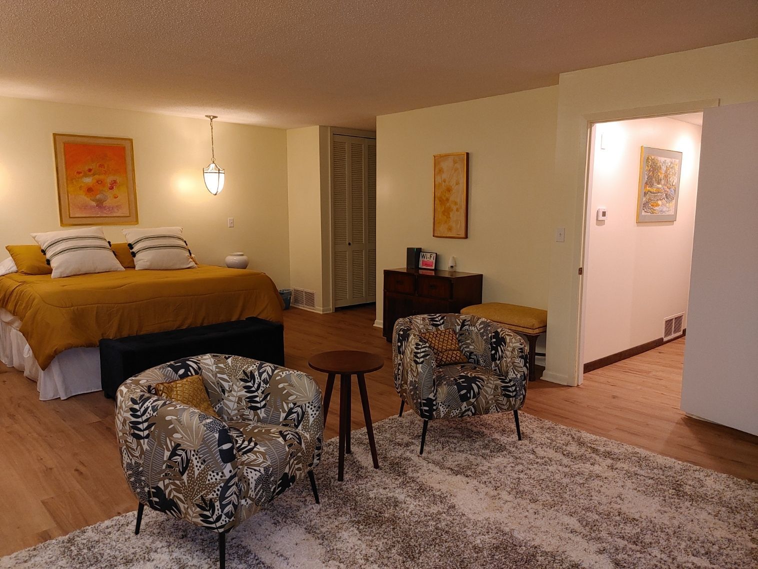 Cozy hotel room with bed, two chairs, small table, rug, and hallway light in the background