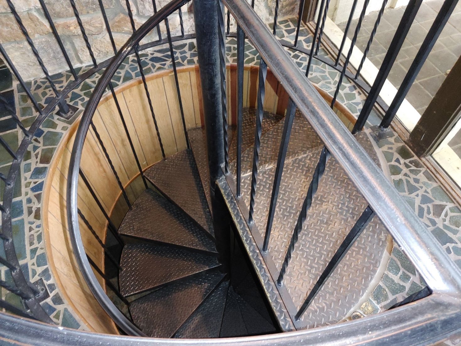 Spiral staircase with black railings and stone-tiled floor, viewed from above.