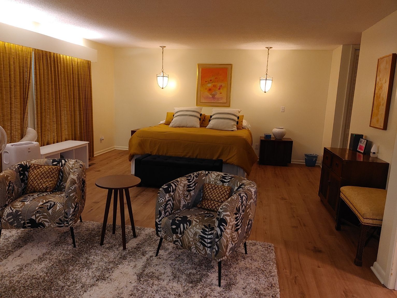 Cozy bedroom with a bed, two armchairs, small stools, warm lighting, and wood floors