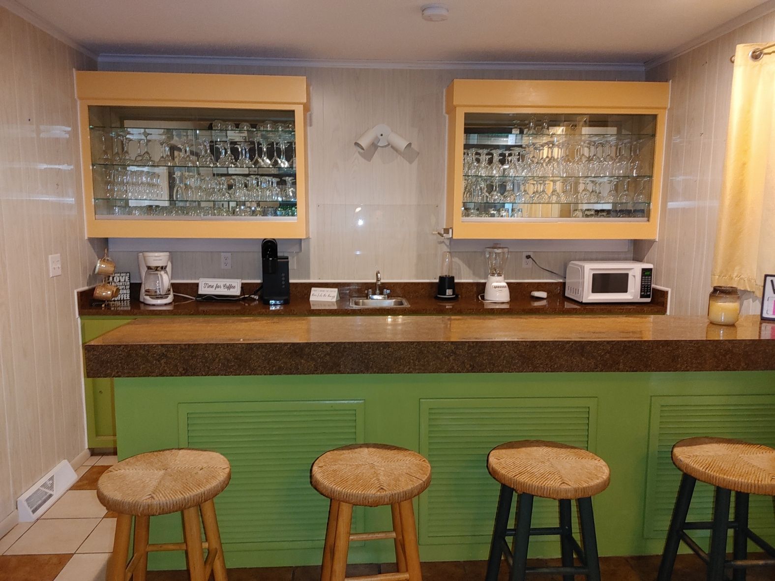 Small bar with green counter, wooden stools, glass cabinets, coffee machine, and microwave in a cozy room