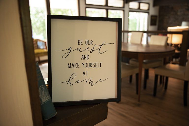 Framed sign on a table reading “Be our guest and make yourself at home” in a cozy room
