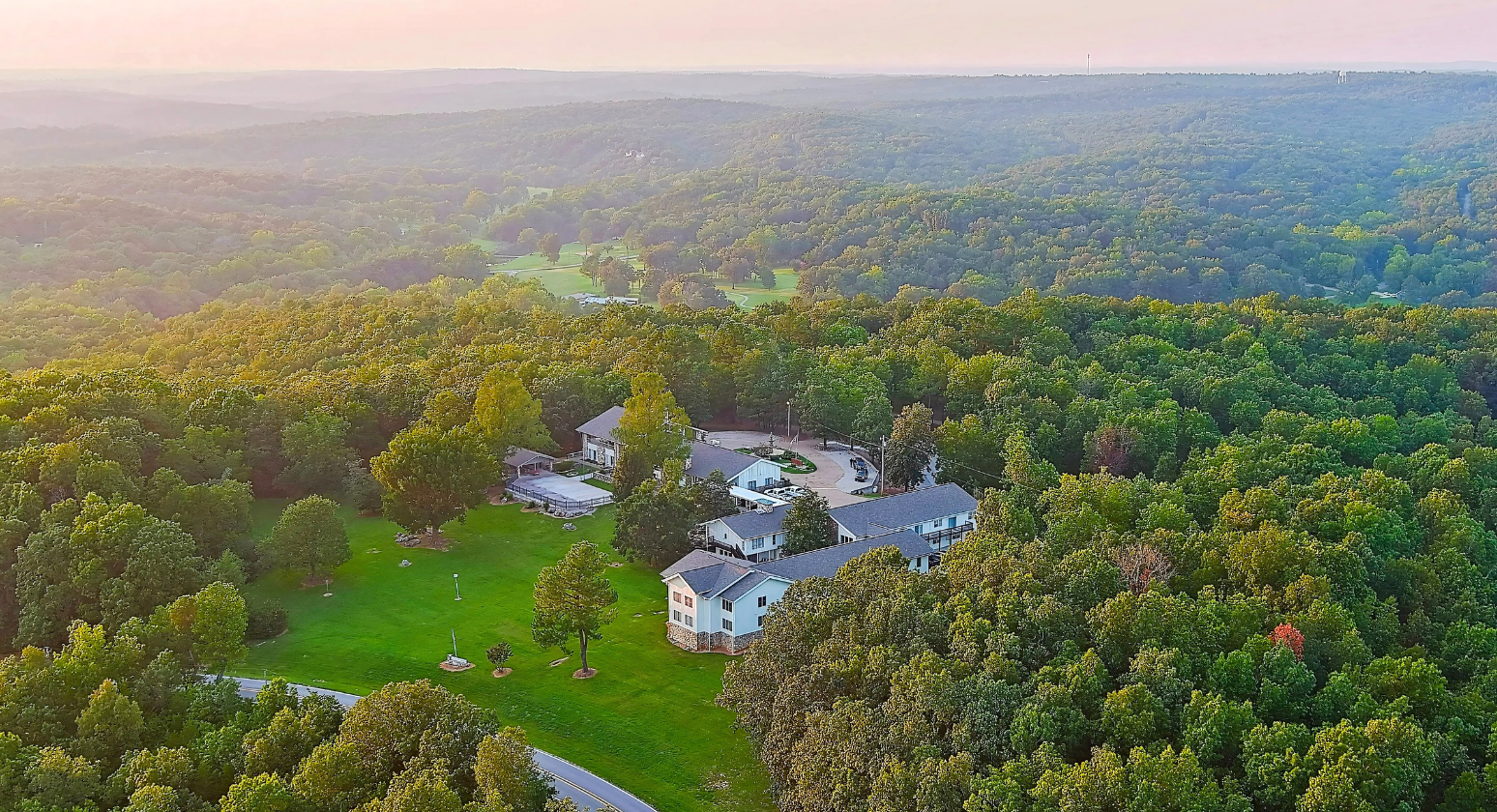 Aerial view of a white house surrounded by lush green forest at sunset.