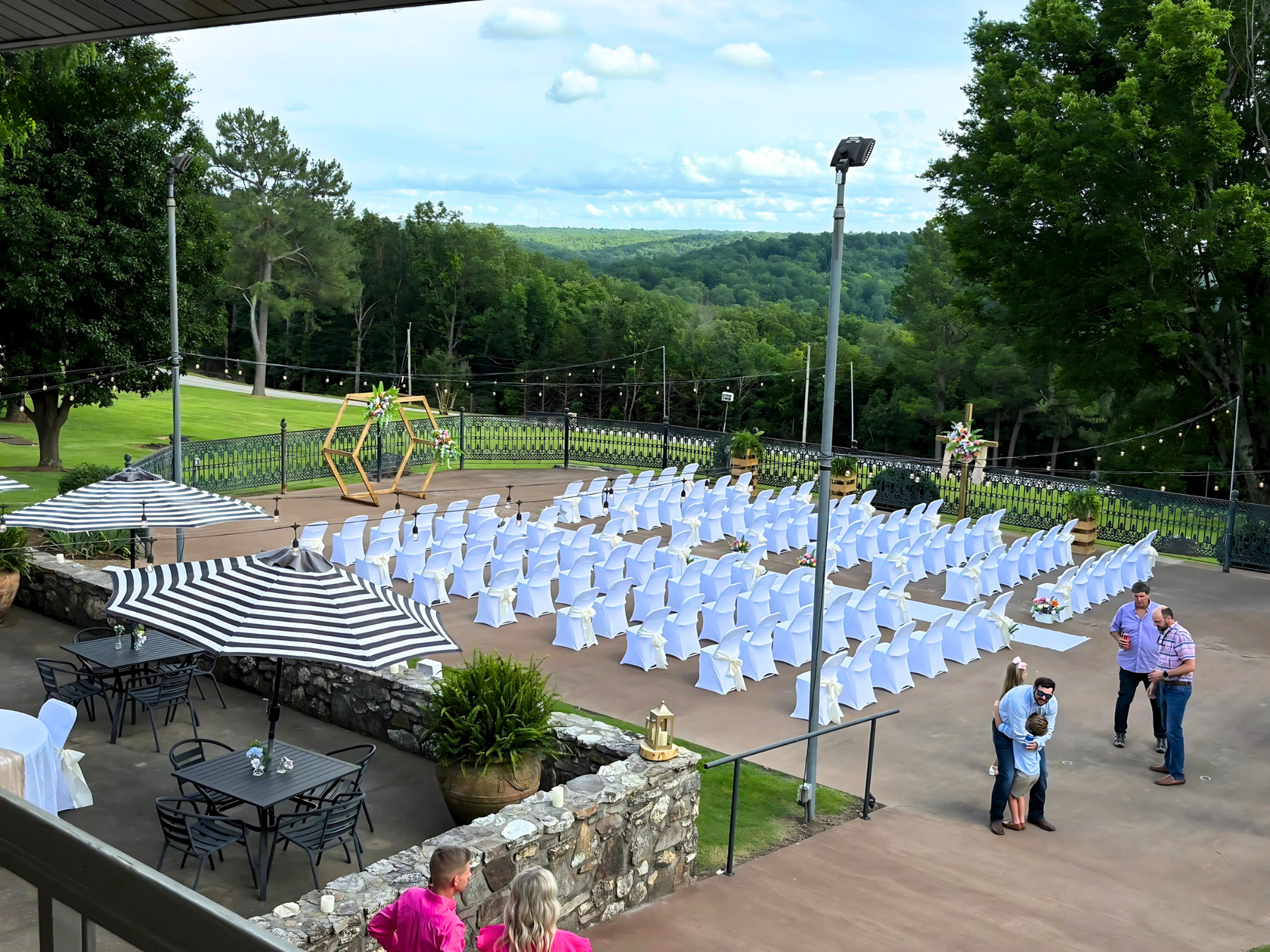 Outdoor wedding setup with rows of white chairs on a patio, striped umbrellas, and two people standing nearby.