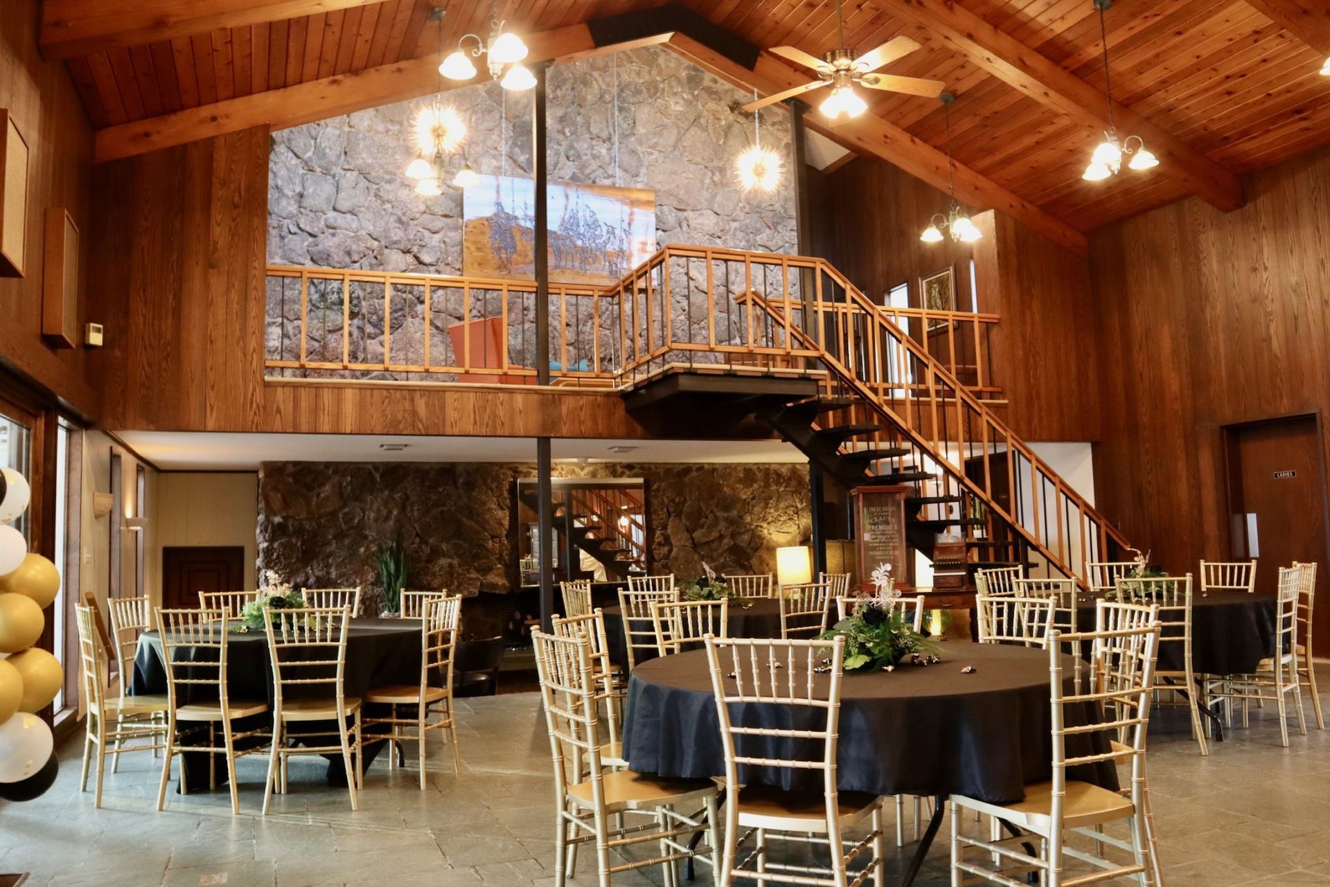 Rustic barn reception hall with round tables, gold chairs, and a loft balcony under wooden beams