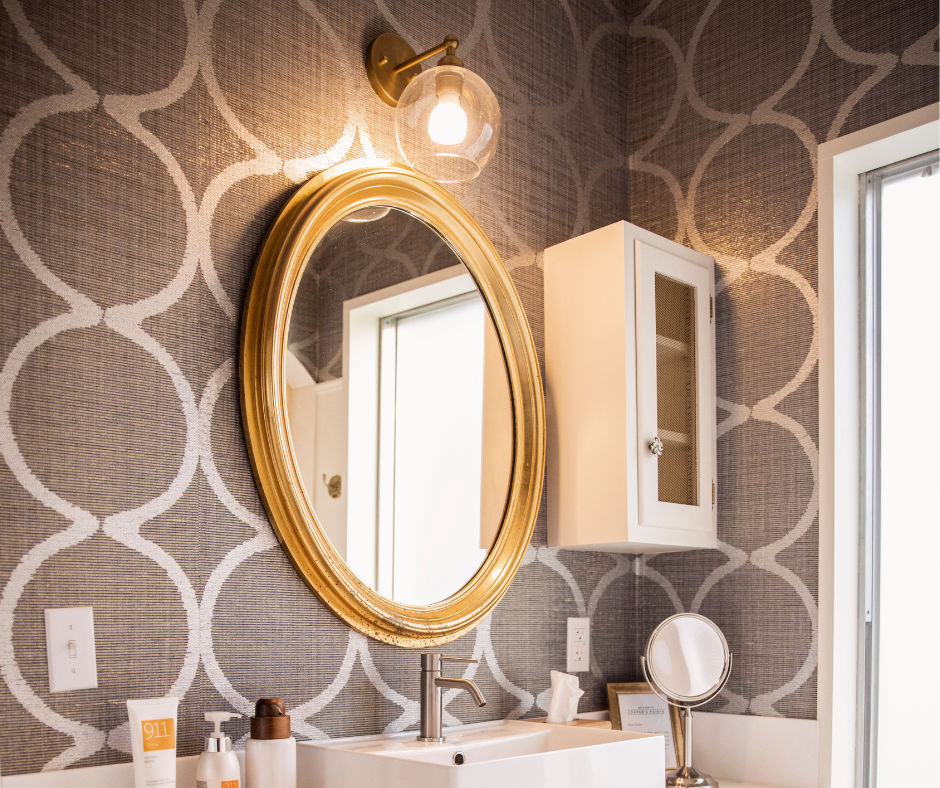 Gold oval mirror above a bathroom sink with patterned wallpaper, vanity lights, and wall cabinet