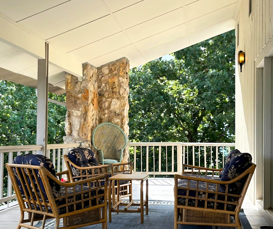 Covered porch with wicker chairs, wooden table, and stone fireplace overlooking a leafy view.