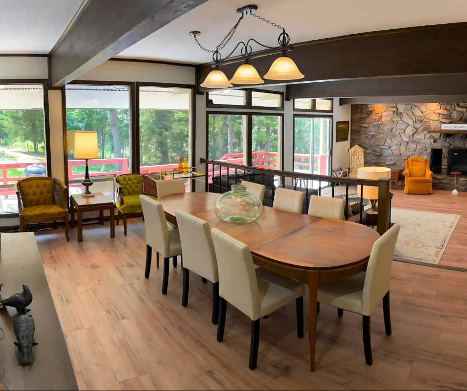 Modern dining room with wooden table, beige chairs, pendant lights, and large windows overlooking greenery