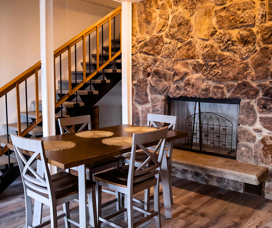 Rustic dining area with wooden table, chairs, stone fireplace, and staircase in the background