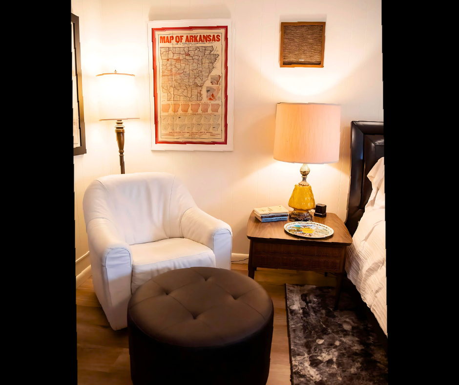 Cozy bedroom corner with white chair, black ottoman, bedside lamp, framed art, and dark wood nightstand