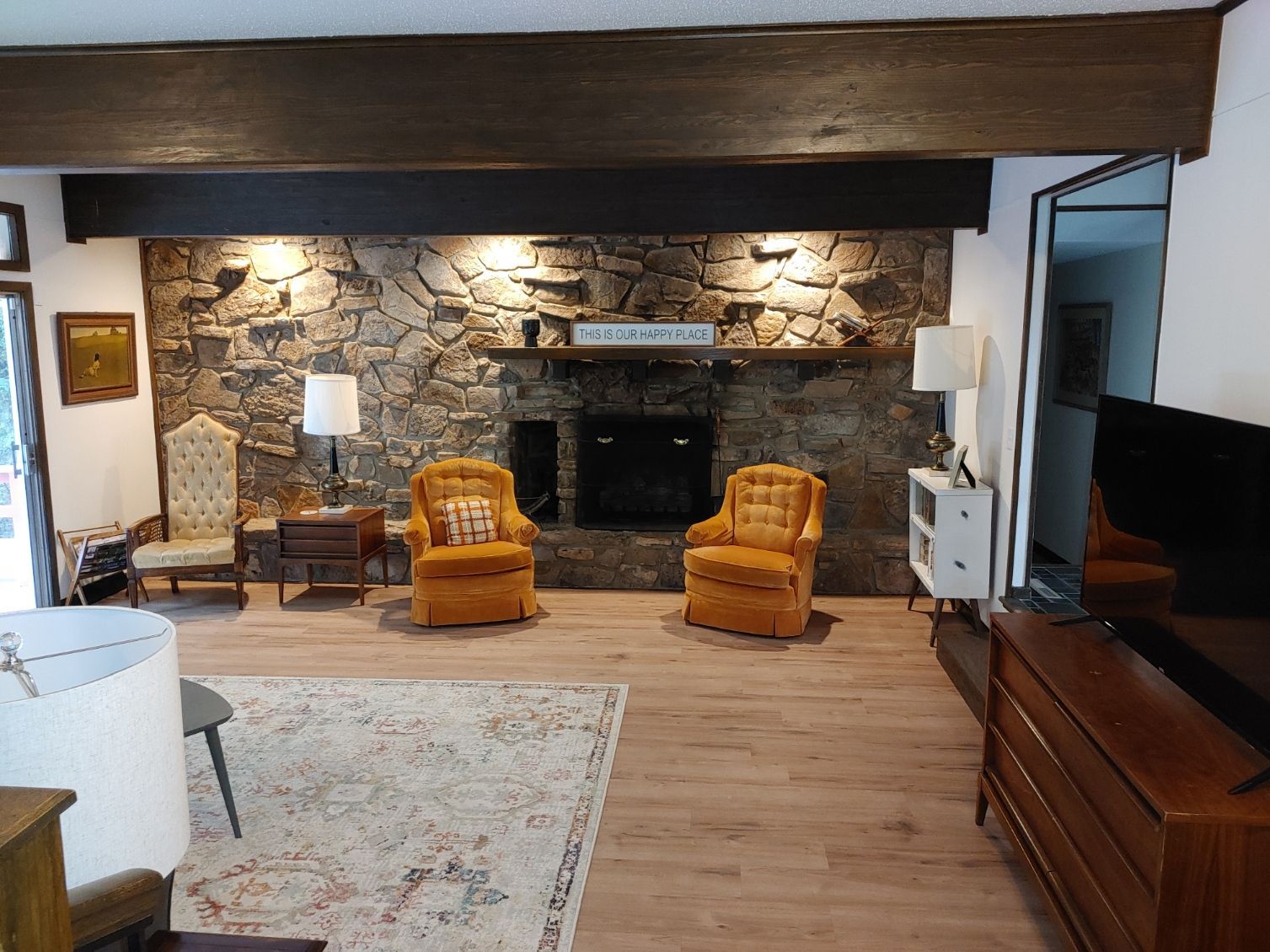 Rustic living room with stone fireplace, two orange chairs, lamps, and hardwood floors