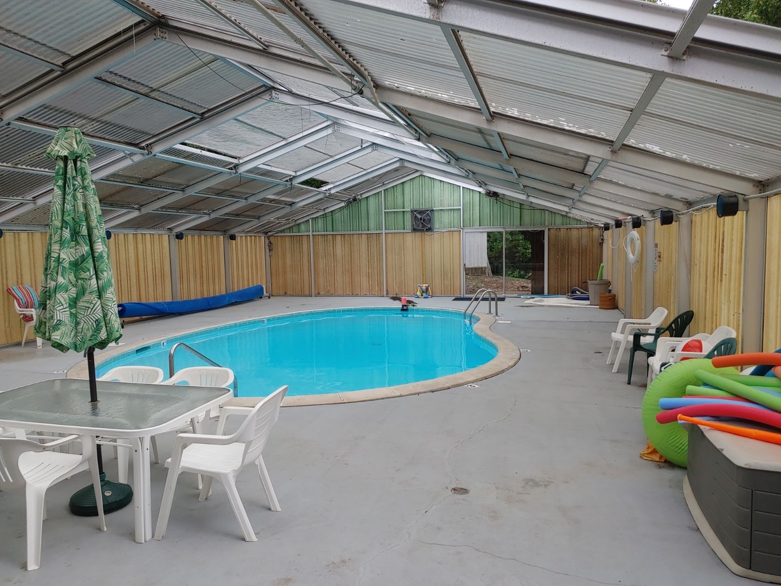 Indoor pool under a metal roof, with white chairs, tables, and colorful pool floats along the side