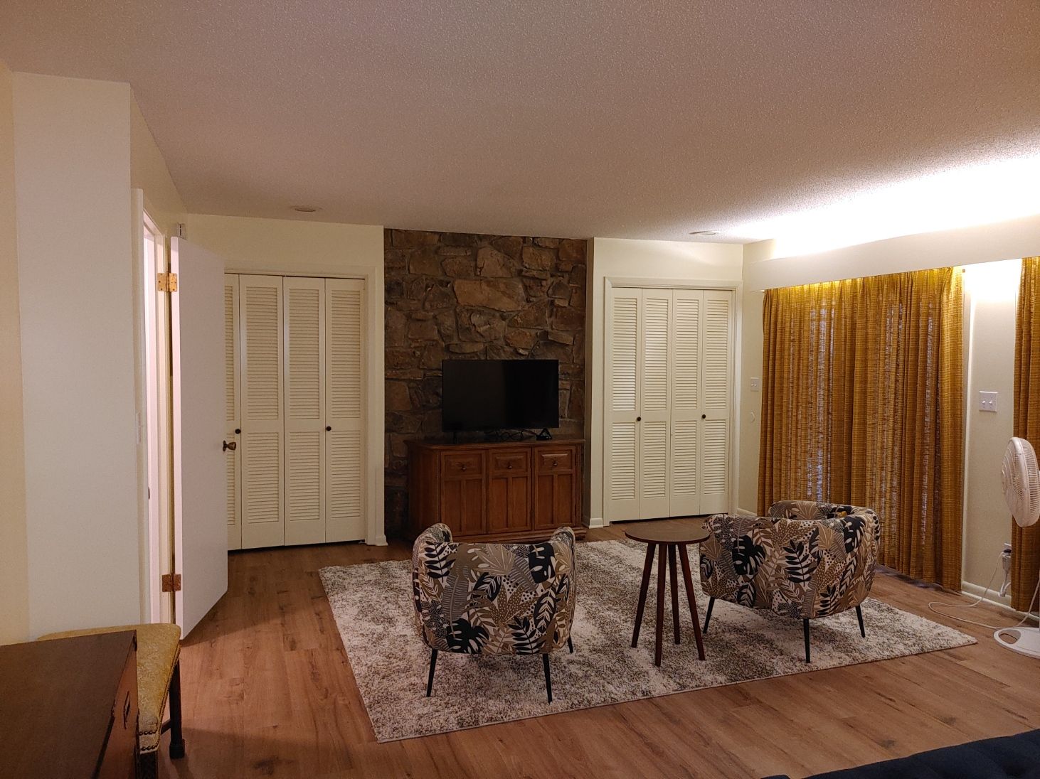 Living room with stone fireplace, TV, white shutters, patterned chairs, and a rug on hardwood floor
