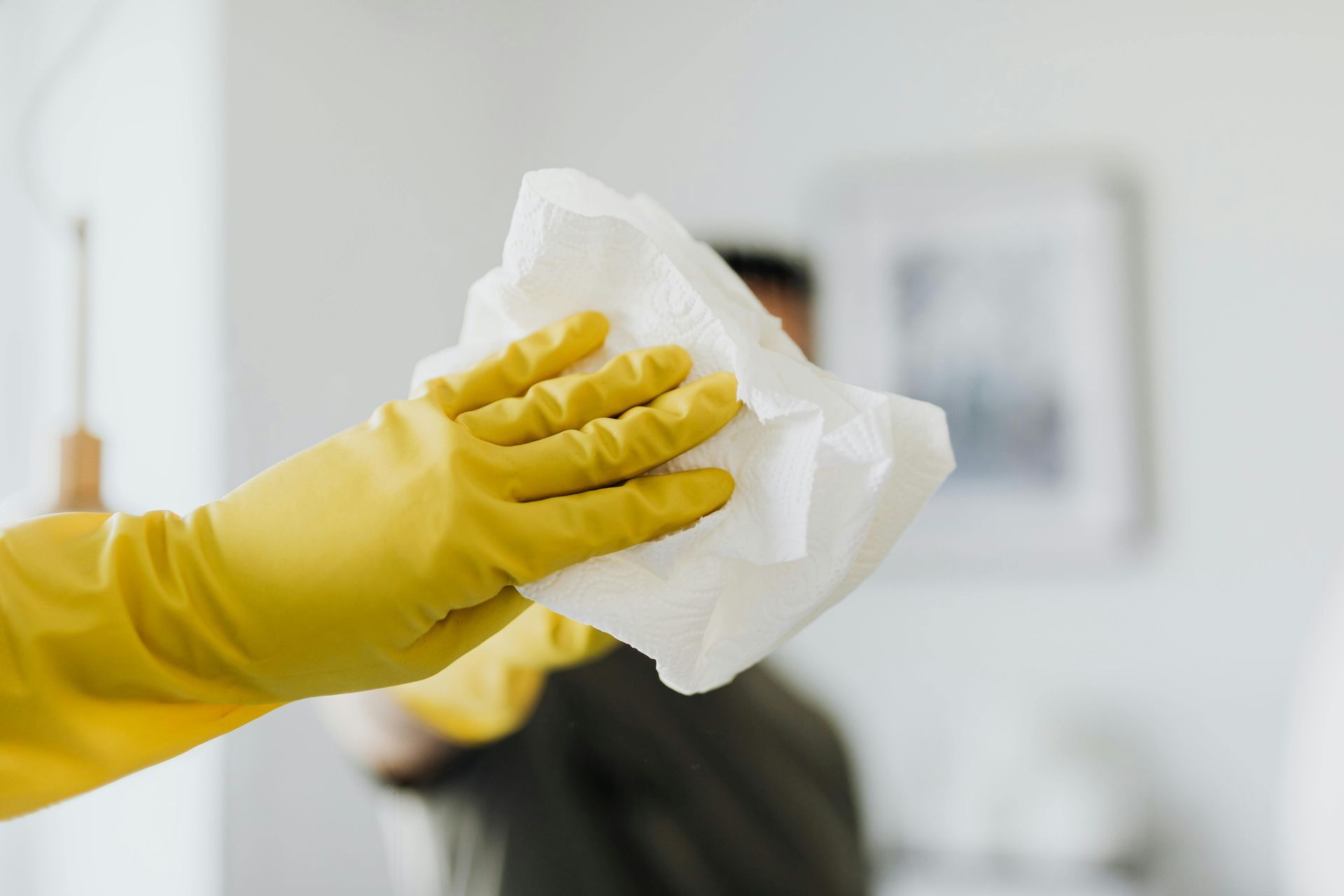A hand wearing a bright yellow cleaning glove holds a white cloth, reaching out toward the foreground.
