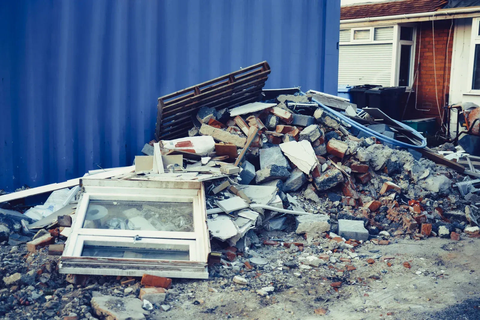 A large pile of construction waste, including a white window frame and bricks, sits against a blue wall near a house.