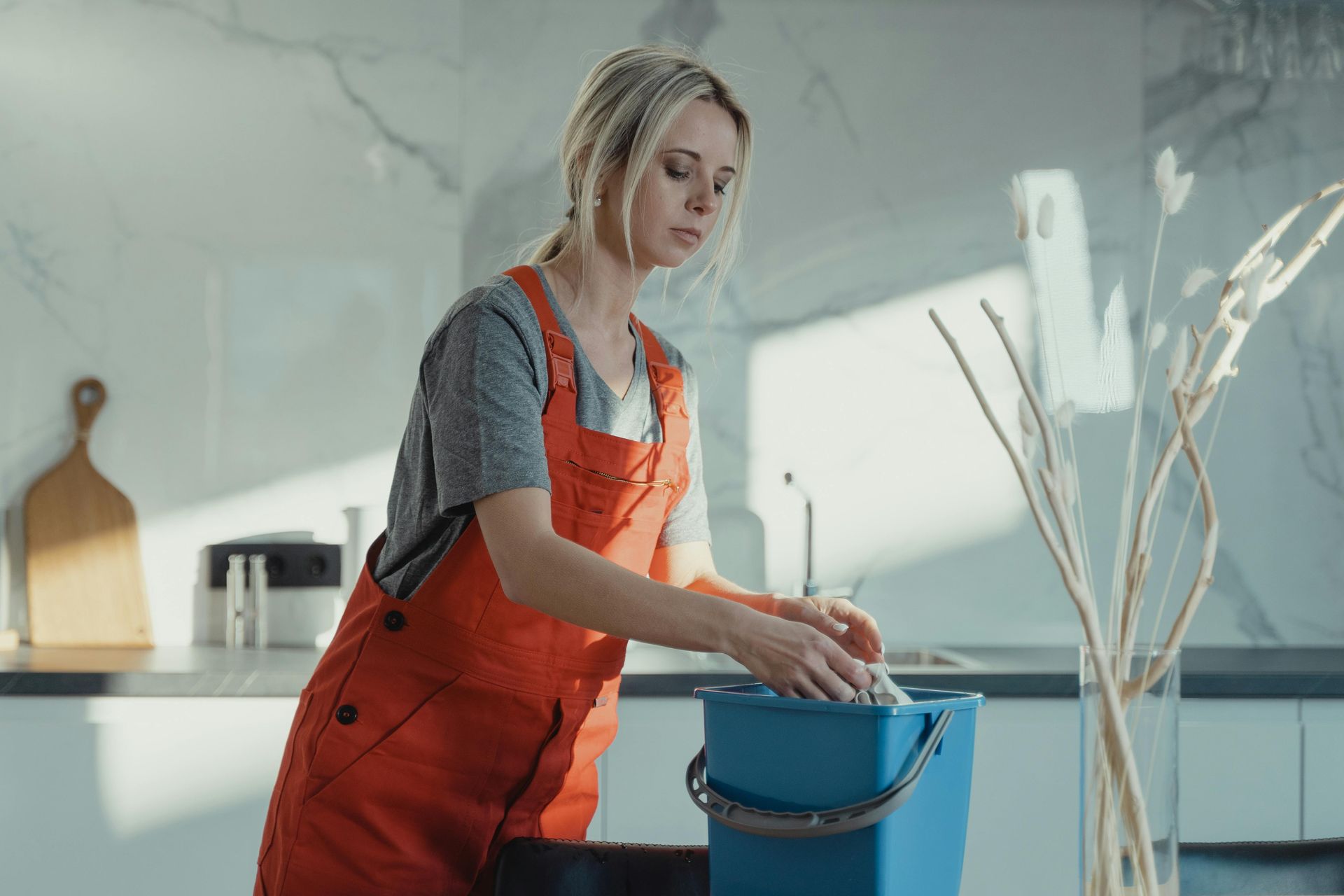 A person in orange overalls cleans a surface while wringing a cloth into a blue bucket in a bright kitchen.