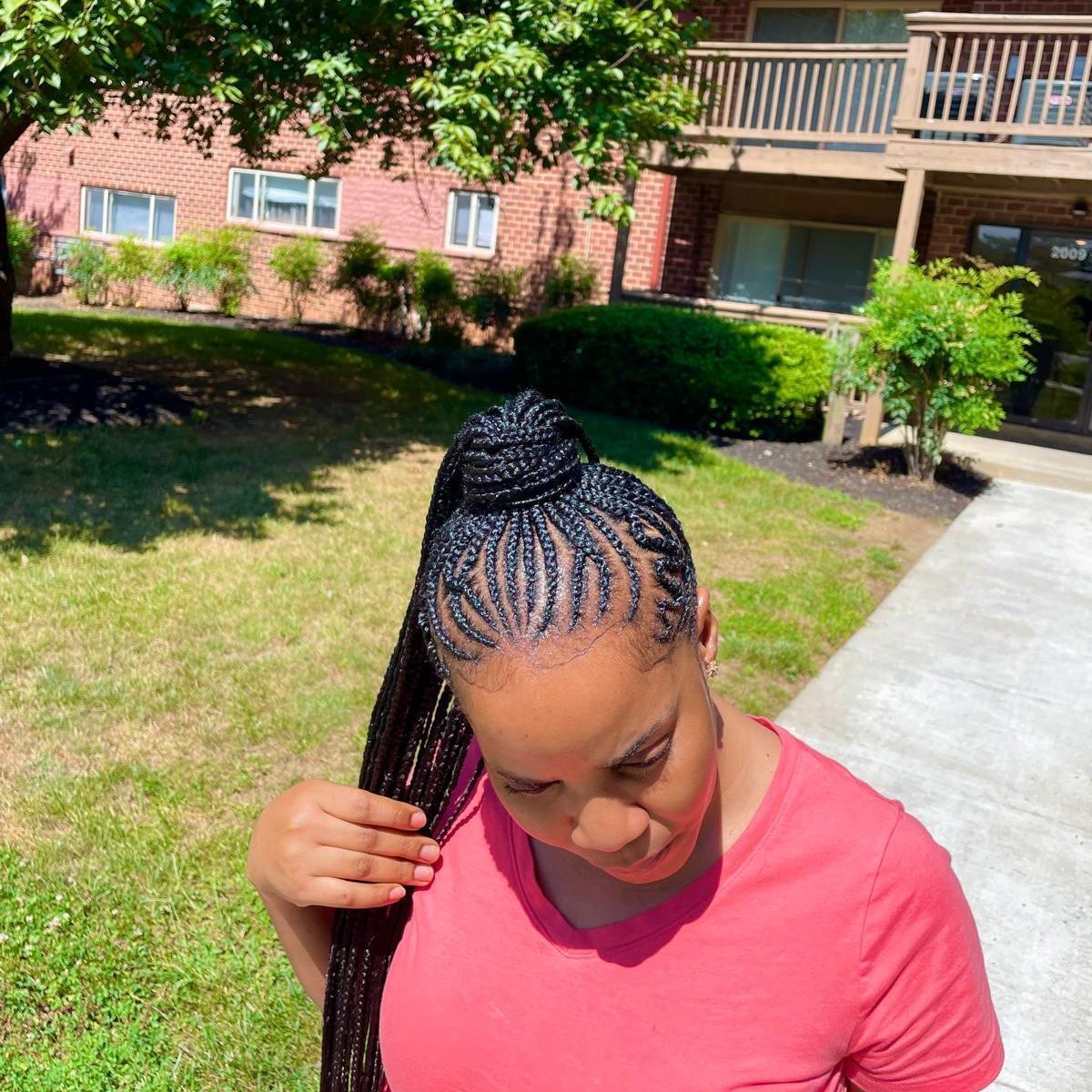 Person with braided hair in a pink shirt standing outdoors near an apartment building.