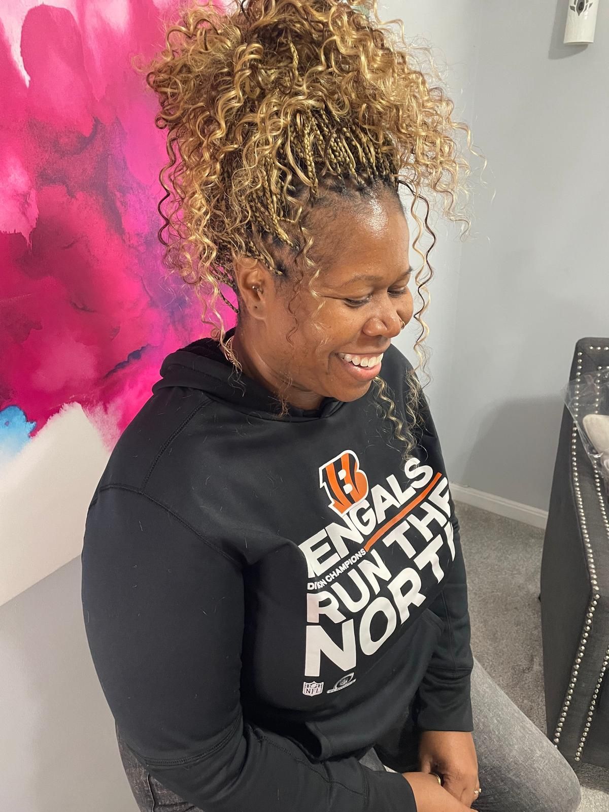 Smiling woman in a black Bengals hoodie seated indoors against a pink wall.