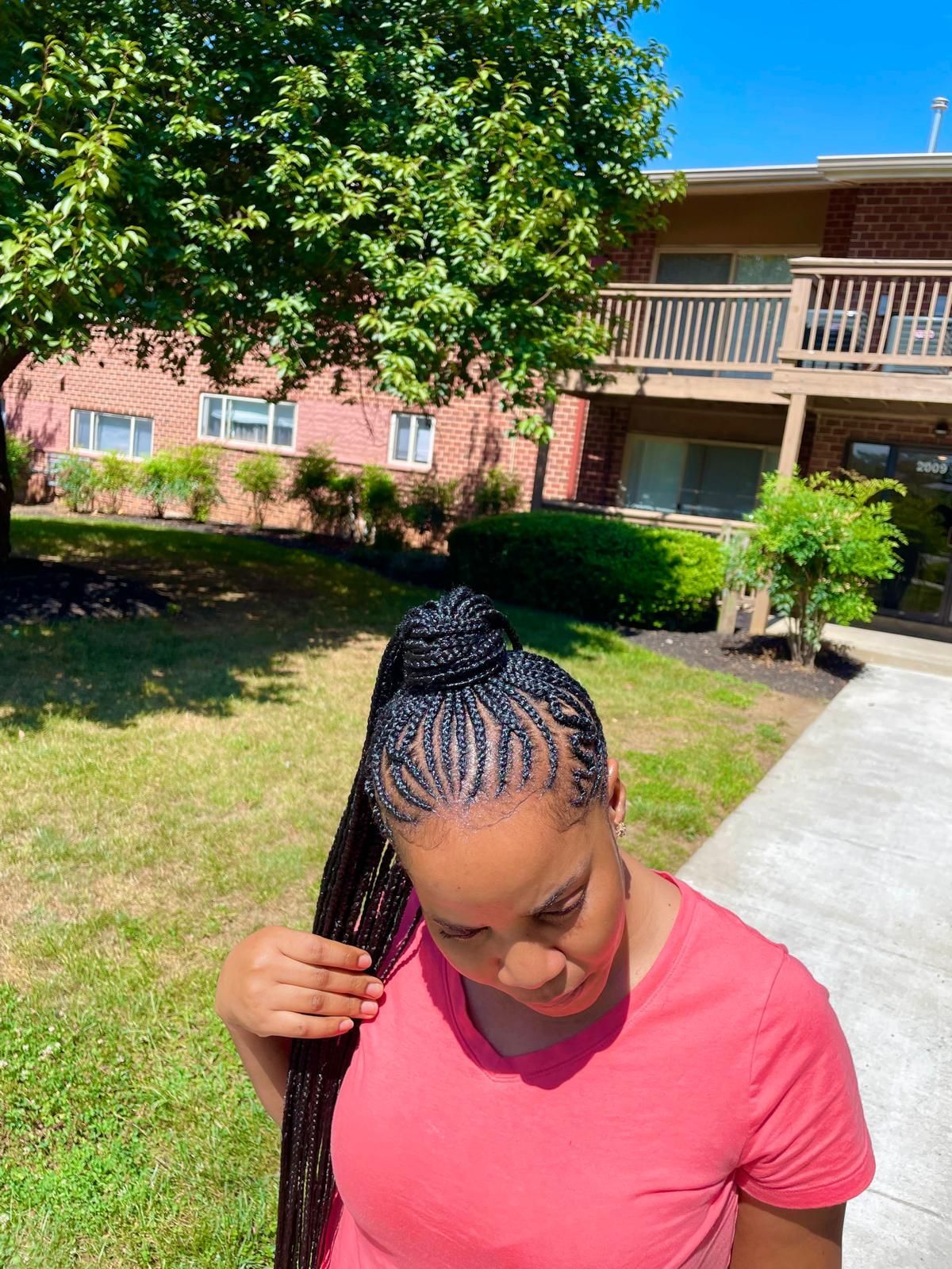 Person with braided hair in a red shirt outdoors near a brick apartment building, looking down.