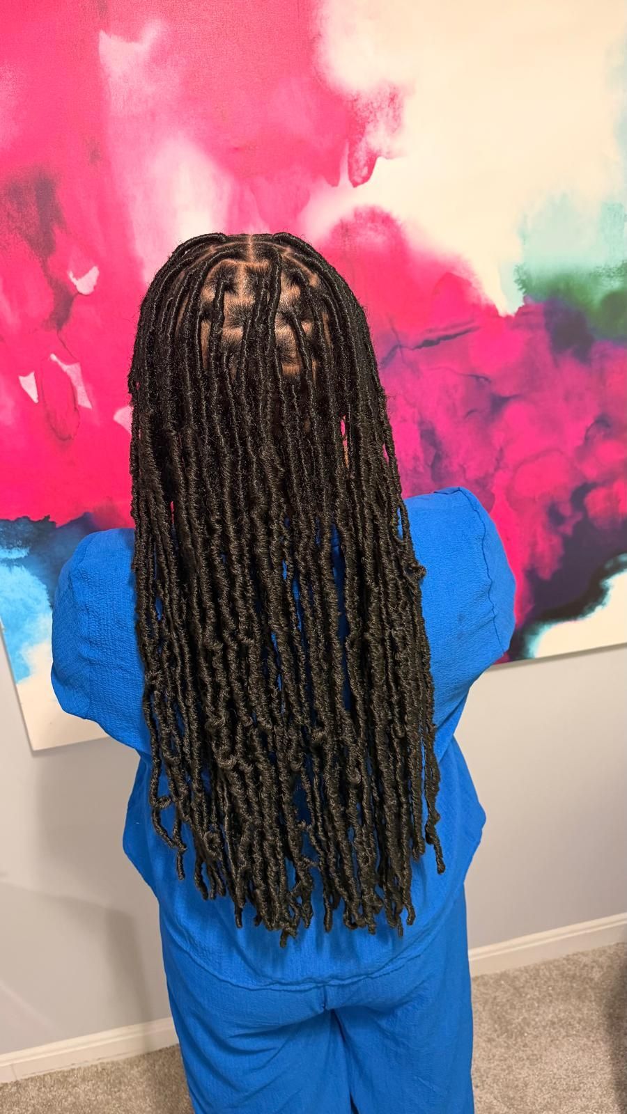 Person in blue outfit facing a colorful abstract painting with long braided hair.