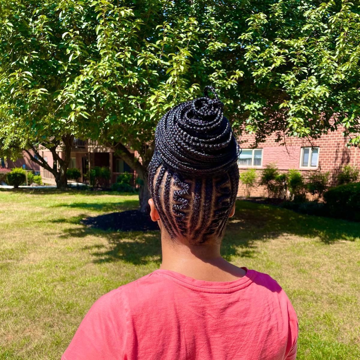 Person with braided bun seen from behind in a pink shirt standing in a sunny park.