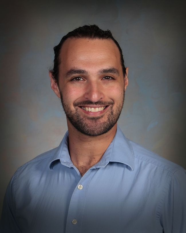 Smiling person in a light blue shirt against a neutral gray studio backdrop