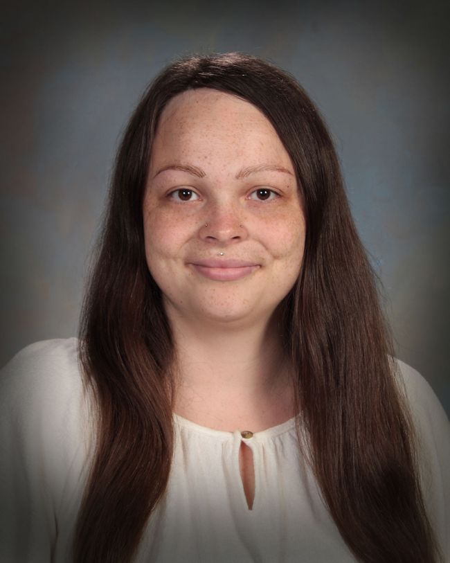 Portrait of a person with long dark hair, wearing a white top, against a gray studio background.