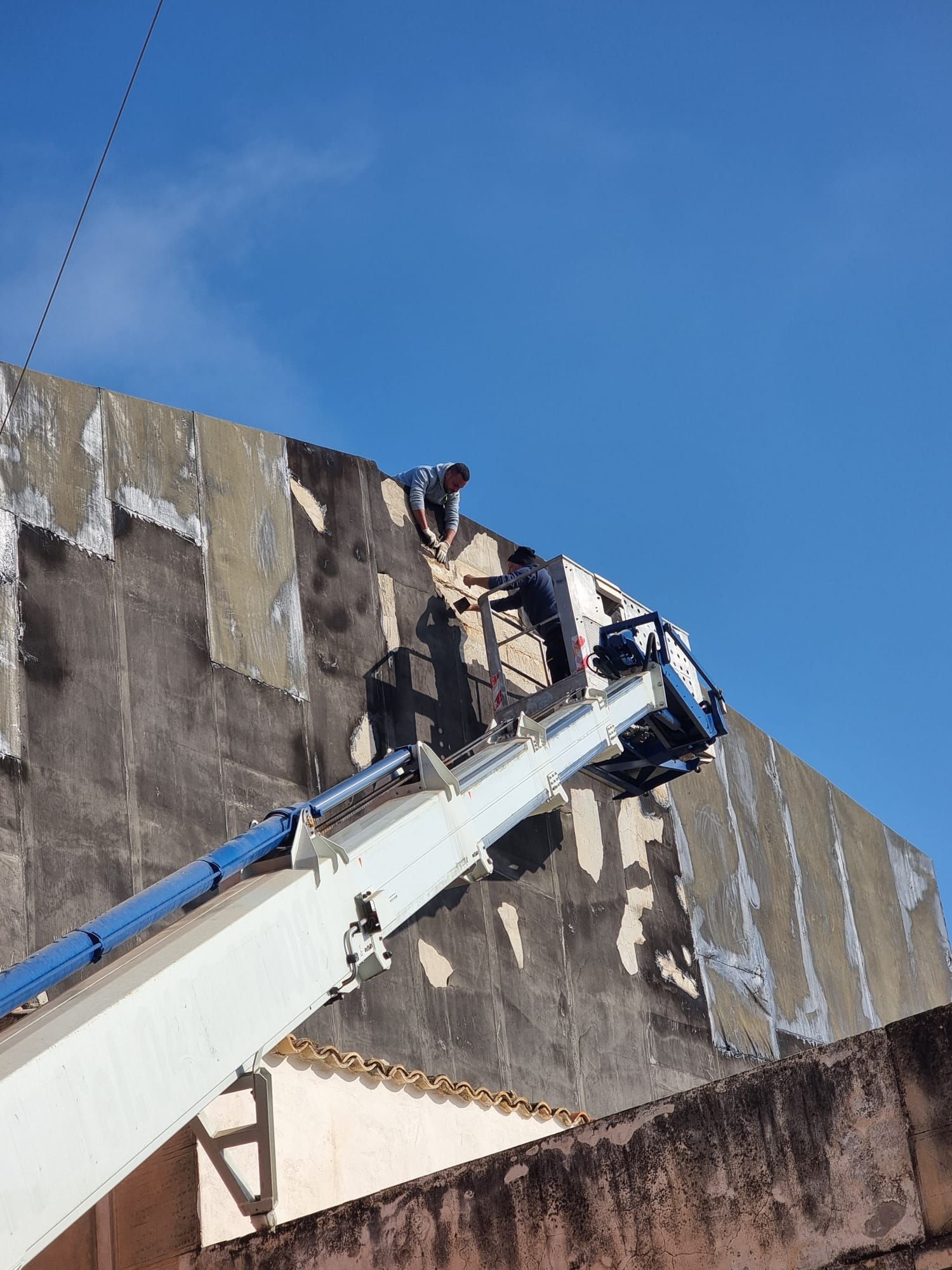 Due operai su una piattaforma aerea stanno lavorando all'esterno di un edificio. Sullo sfondo il cielo azzurro.