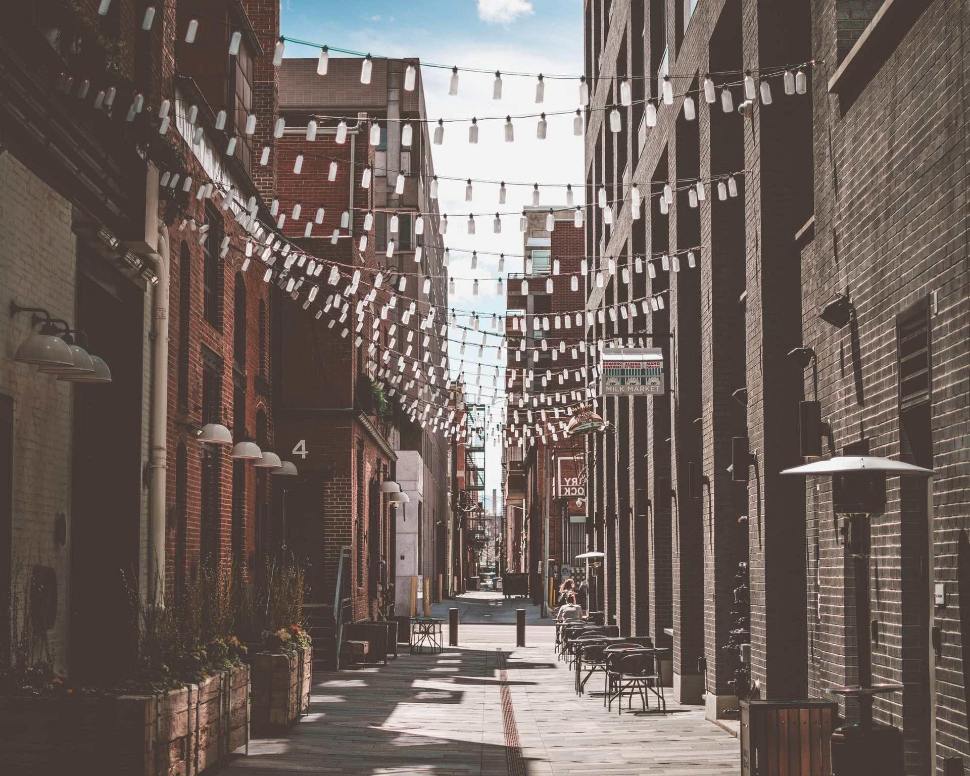 Narrow brick alley with hanging string lights, tables, and chairs in warm sunlight